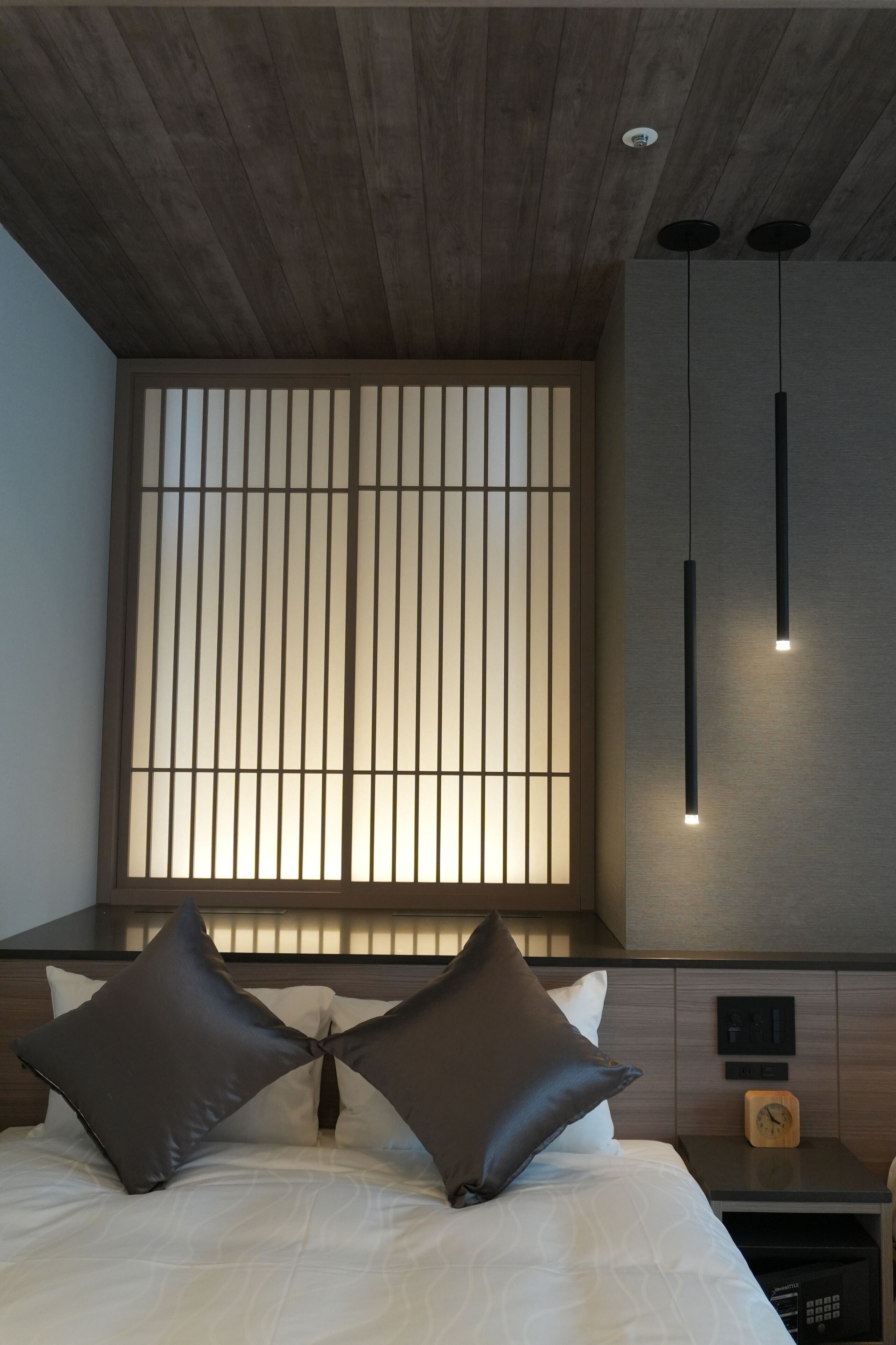 Ein modernes Hotelzimmer mit Holzboden, einem Bett mit weißer Bettwäsche und zwei dunklen Kissen, einem Nachttisch mit einer Uhr und einer wandmontierten Lampe.