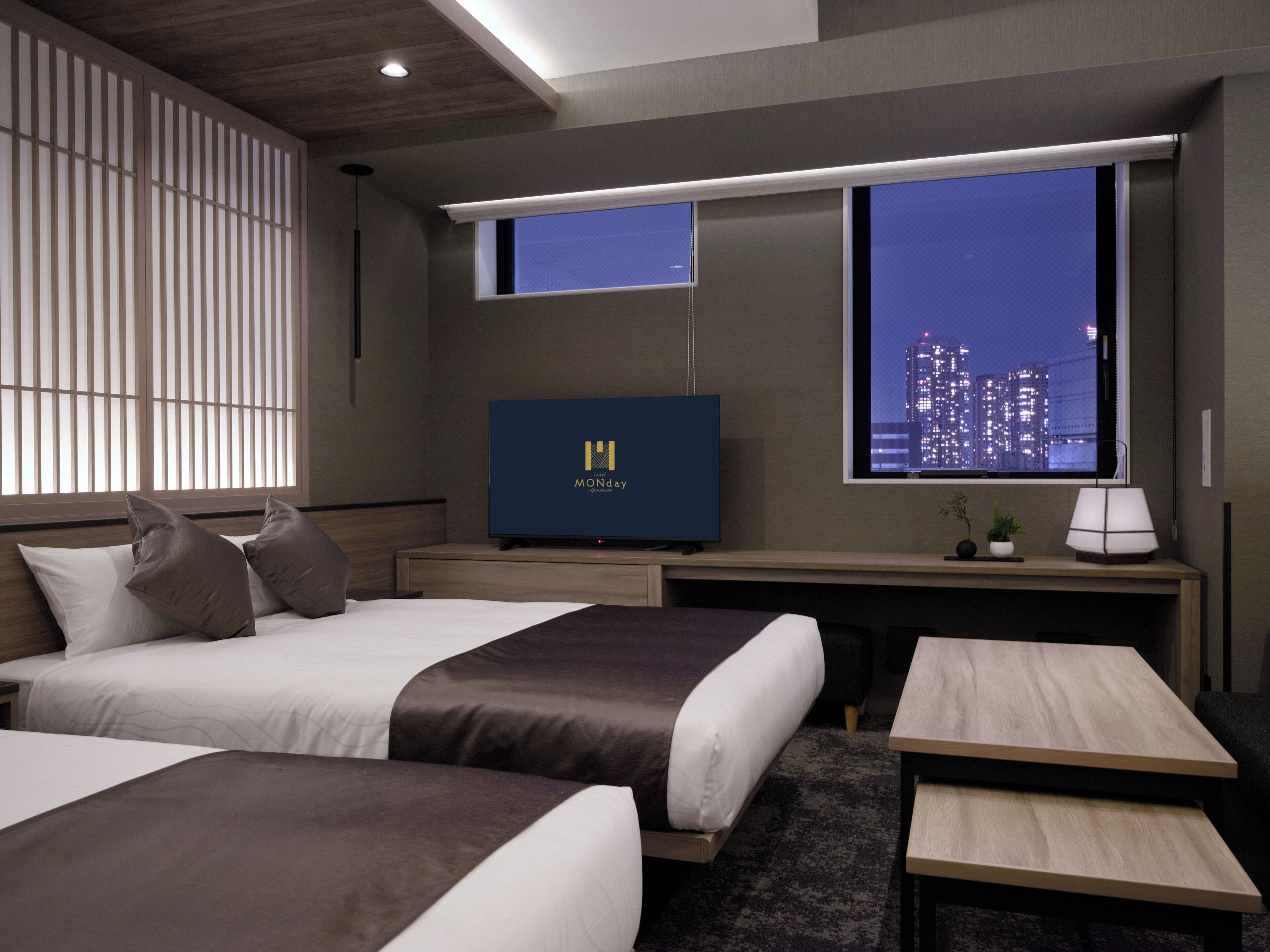 A hotel room with two beds, a desk, a television, and a window with a city view.