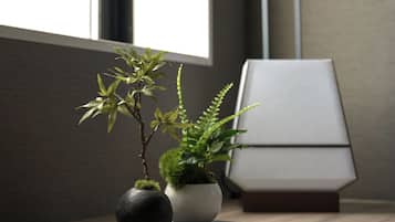 Une petite composition florale en pot, placée sur une surface en bois près d’une fenêtre, avec une lampe moderne et un livre en arrière-plan.