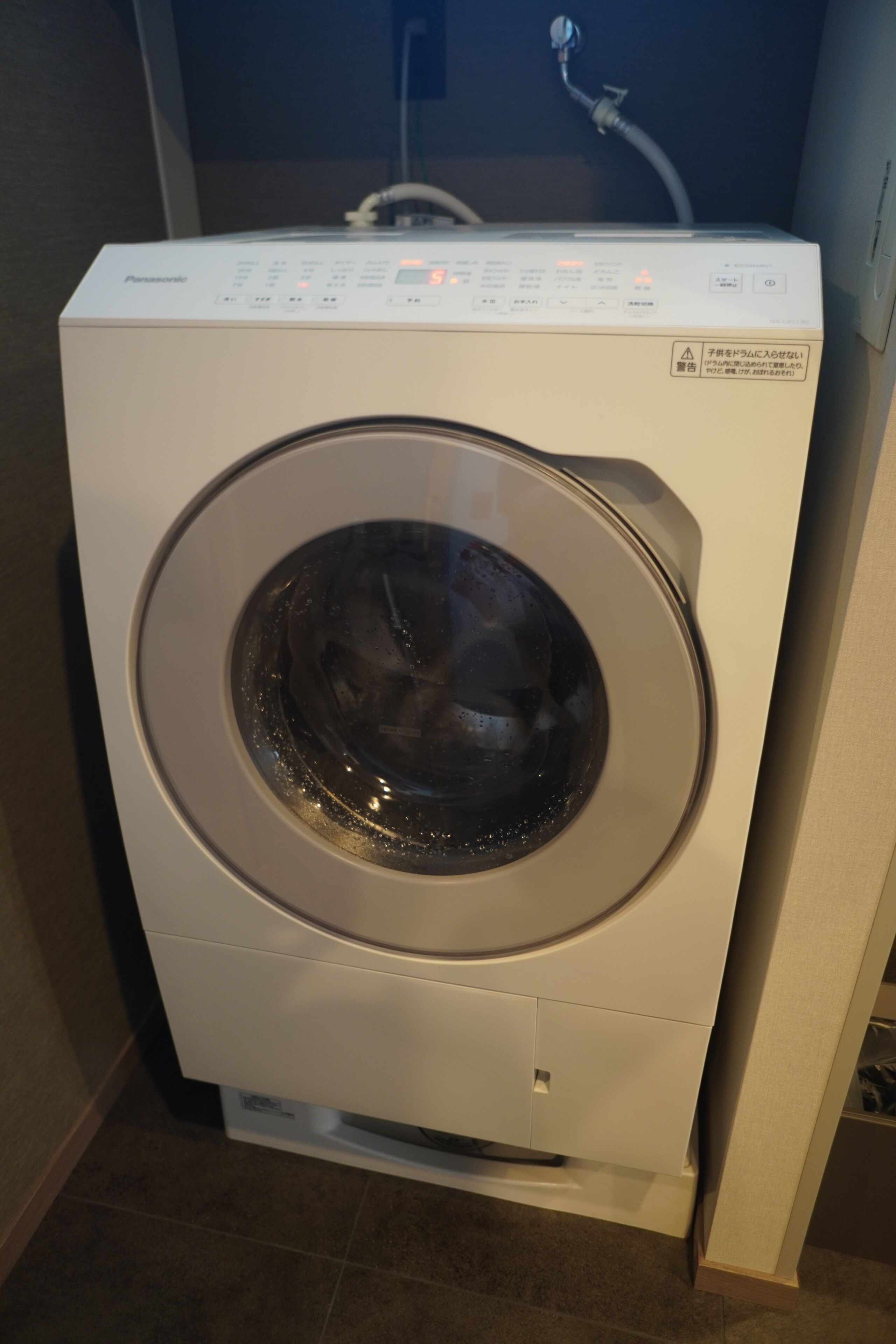 Eine Waschmaschine mit Frontlader in einem Raum mit Fliesenboden und einer sichtbaren Sanitärinstallation im Hintergrund.
