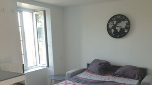 A bedroom with a bed, a window, a bookshelf, and a wall clock. - Furnished studio (Montaigu-Vendée)