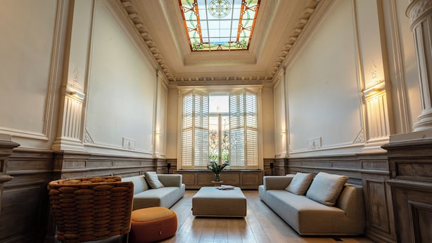 A spacious room with a high ceiling, a large window with shutters, a central ottoman, and two sofas. - Villa Euphrosina (Ostend)