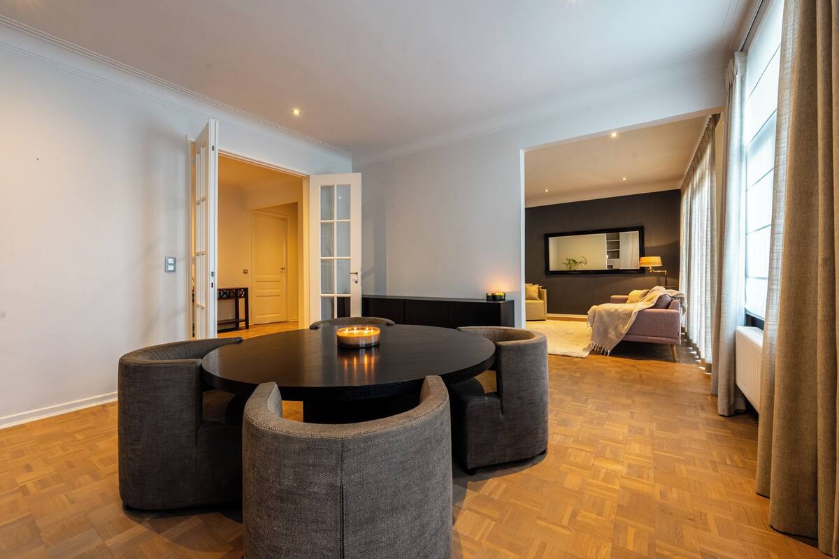 Un salon moderne avec une table ronde noire, des fauteuils recouverts d’un tissu gris, un canapé et une grande fenêtre avec des rideaux.