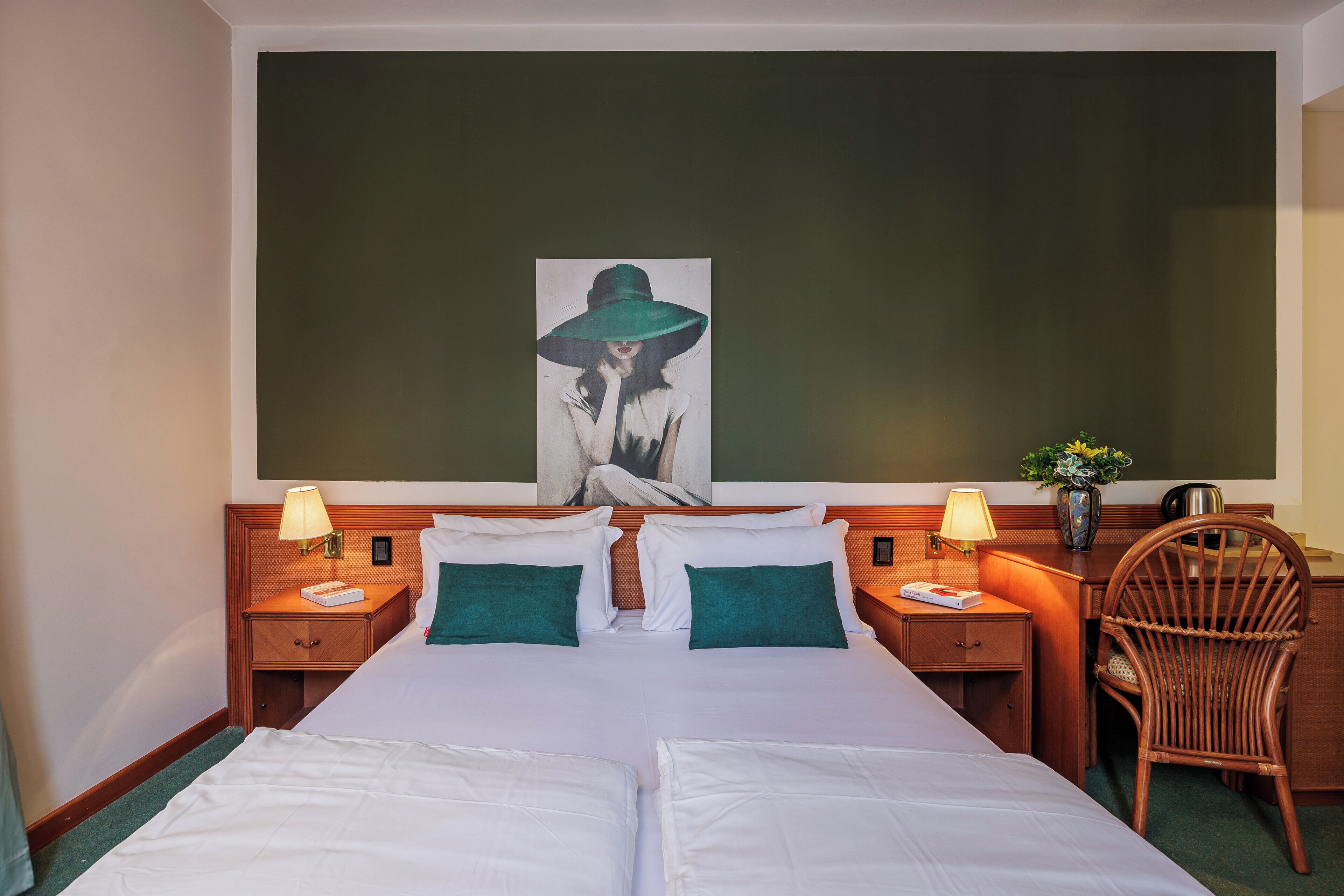 A hotel room with a large bed, two bedside tables with lamps, a wicker chair, and a painting on the wall.