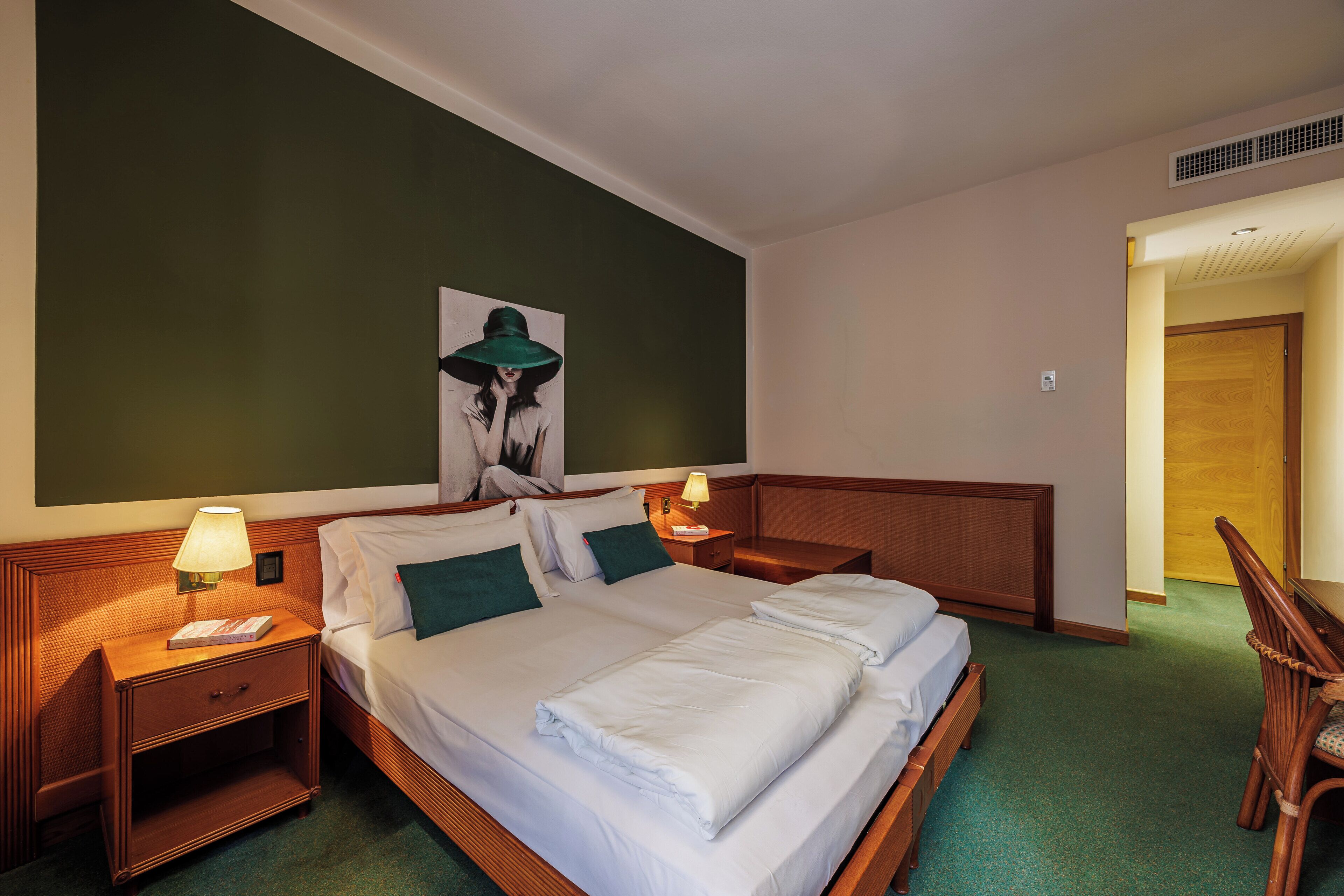 A hotel room with a large bed, two bedside tables with lamps, a wooden nightstand, a chair, and a painting on the wall.