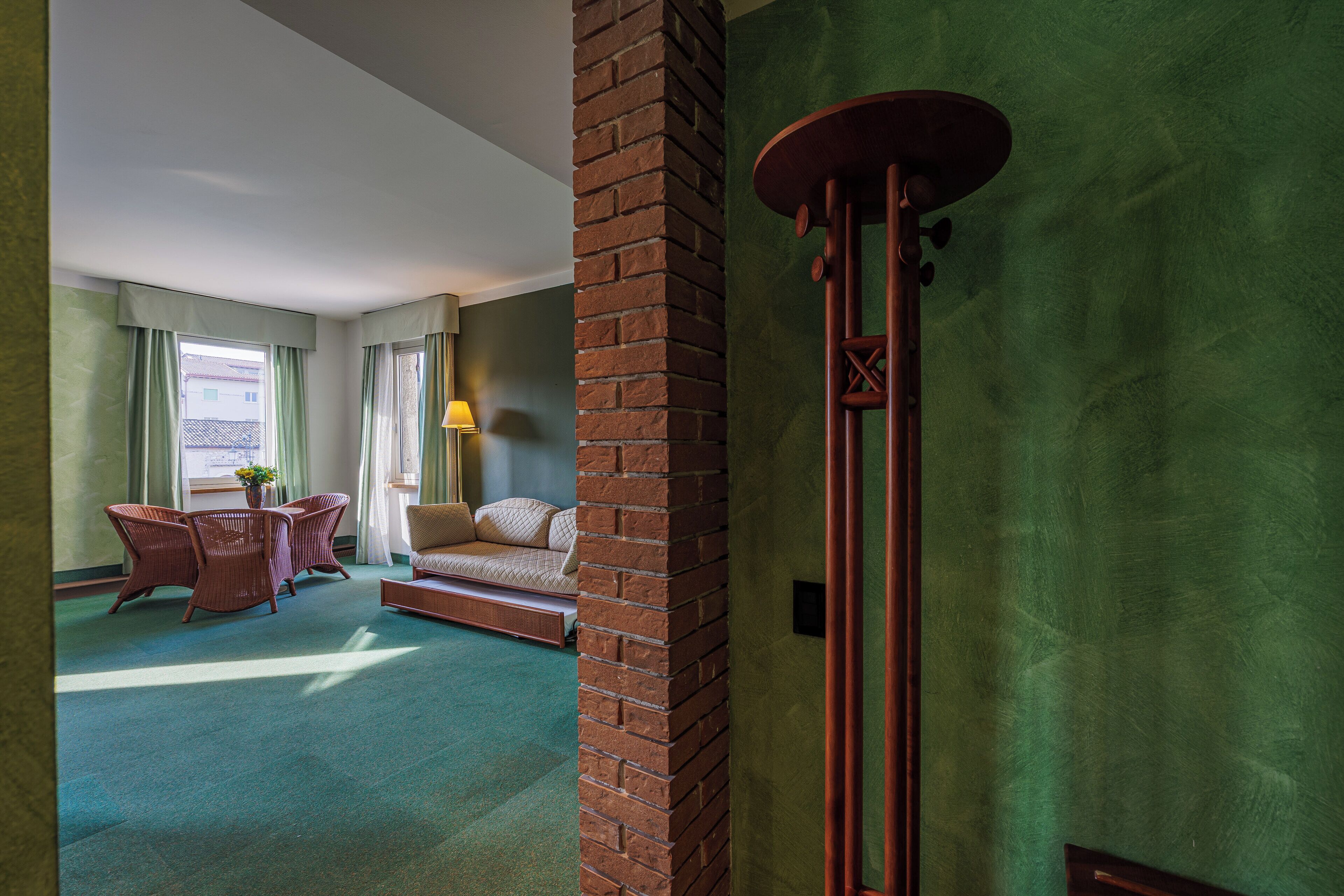 A hotel room with a green carpet, a brick pillar, a wooden chair, a sofa, and a table with a lamp.