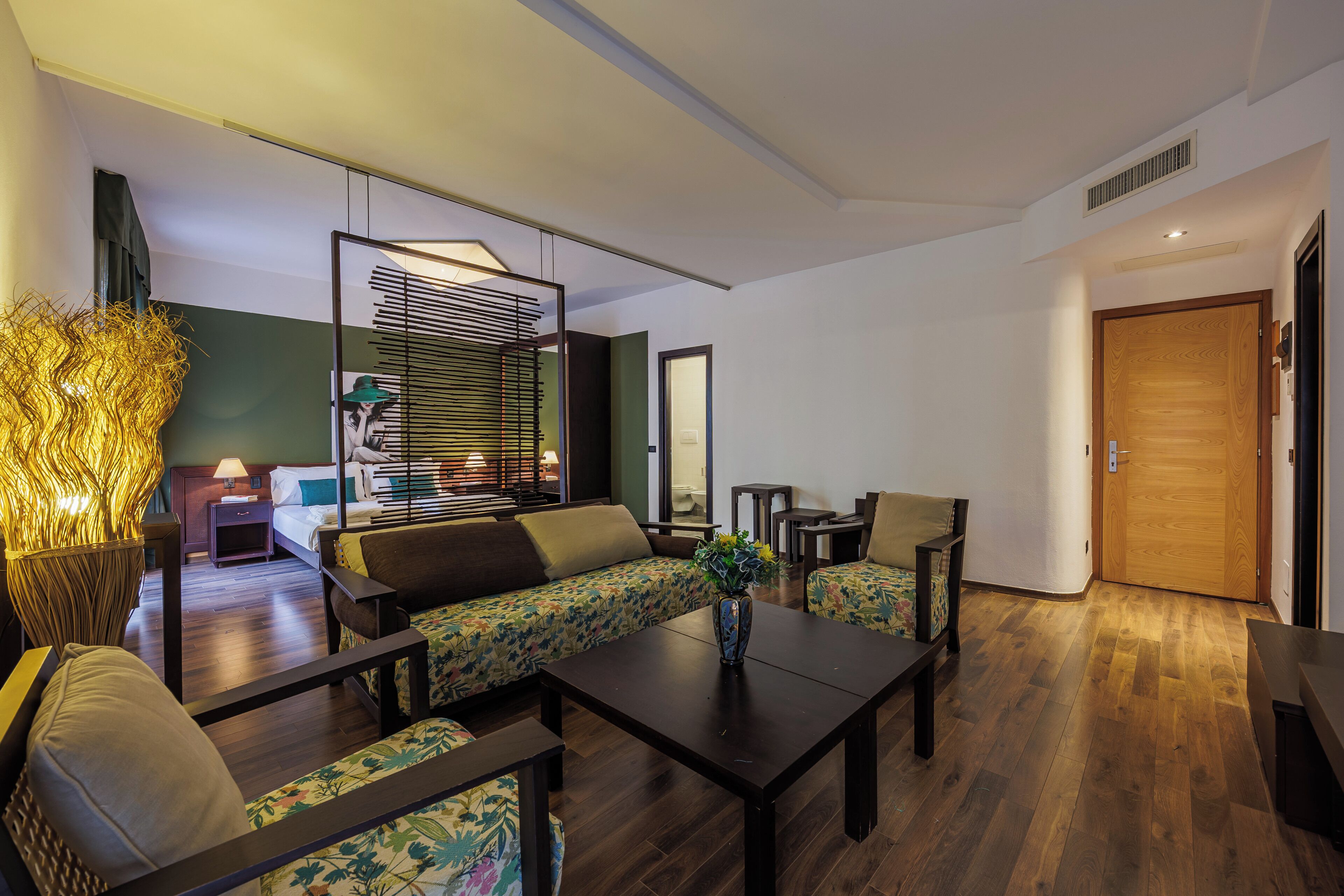 A modern hotel room with a sofa, armchairs, a coffee table, and a large mirror.