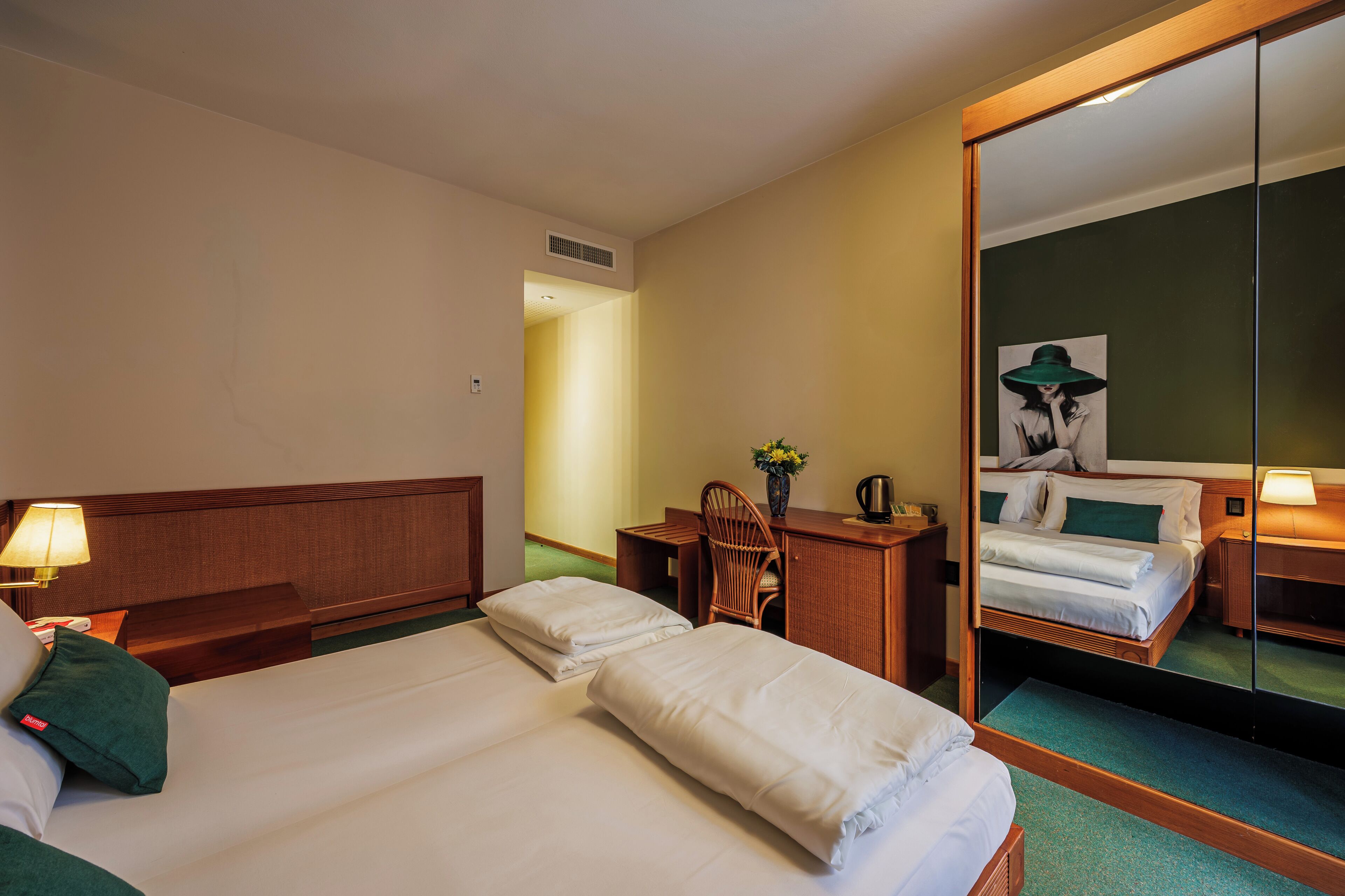 A hotel room with two beds, a desk with a chair, a mirror, and a painting on the wall.