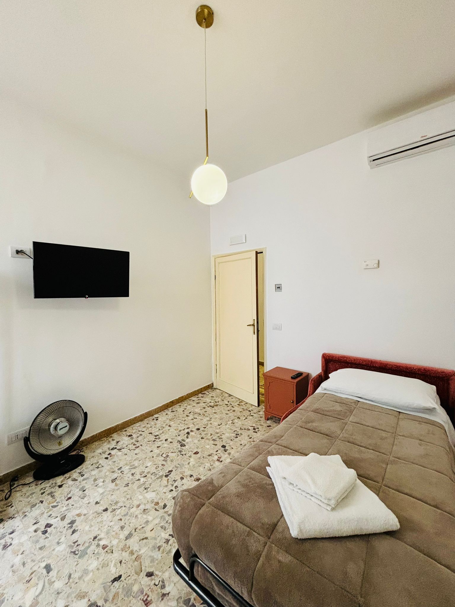 A bedroom with a bed, a television mounted on the wall, a standing fan, and a door leading to another room.