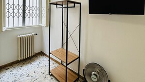 Una habitación con un radiador, un ventilador de pie, un televisor montado en la pared y una ventana con un diseño geométrico.