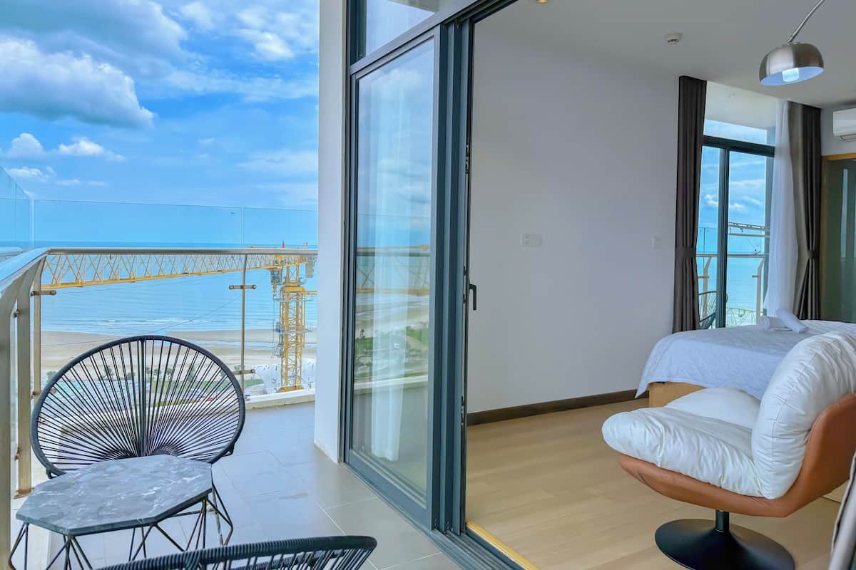 A balcony with a view of the sea and a construction crane, a bed with white bedding, and a black chair.