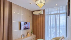 Een moderne woonkamer met een flatscreen televisie bevestigd aan een houten wandpaneel, een luchtverwarmer die aan het plafond is gemonteerd, en een groot raam met dunne gordijnen.