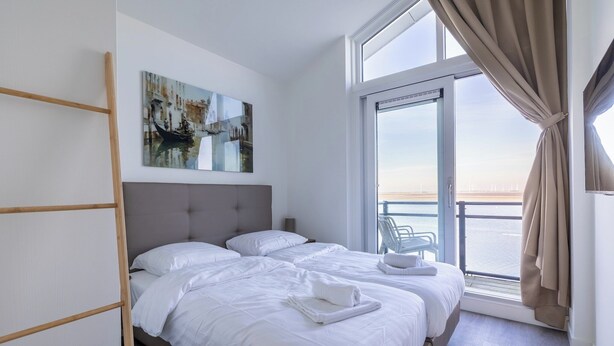Ein modernes Hotelzimmer mit großem Fenster, einem Bett mit weißer Bettwäsche, einem Gemälde an der Wand und einem Balkon mit Meerblick.