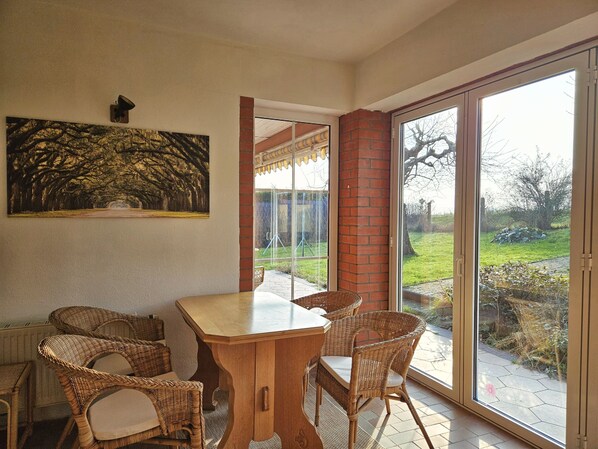 A dining area with a wooden table and wicker chairs, a large painting on the wall, and a glass door leading to a garden.