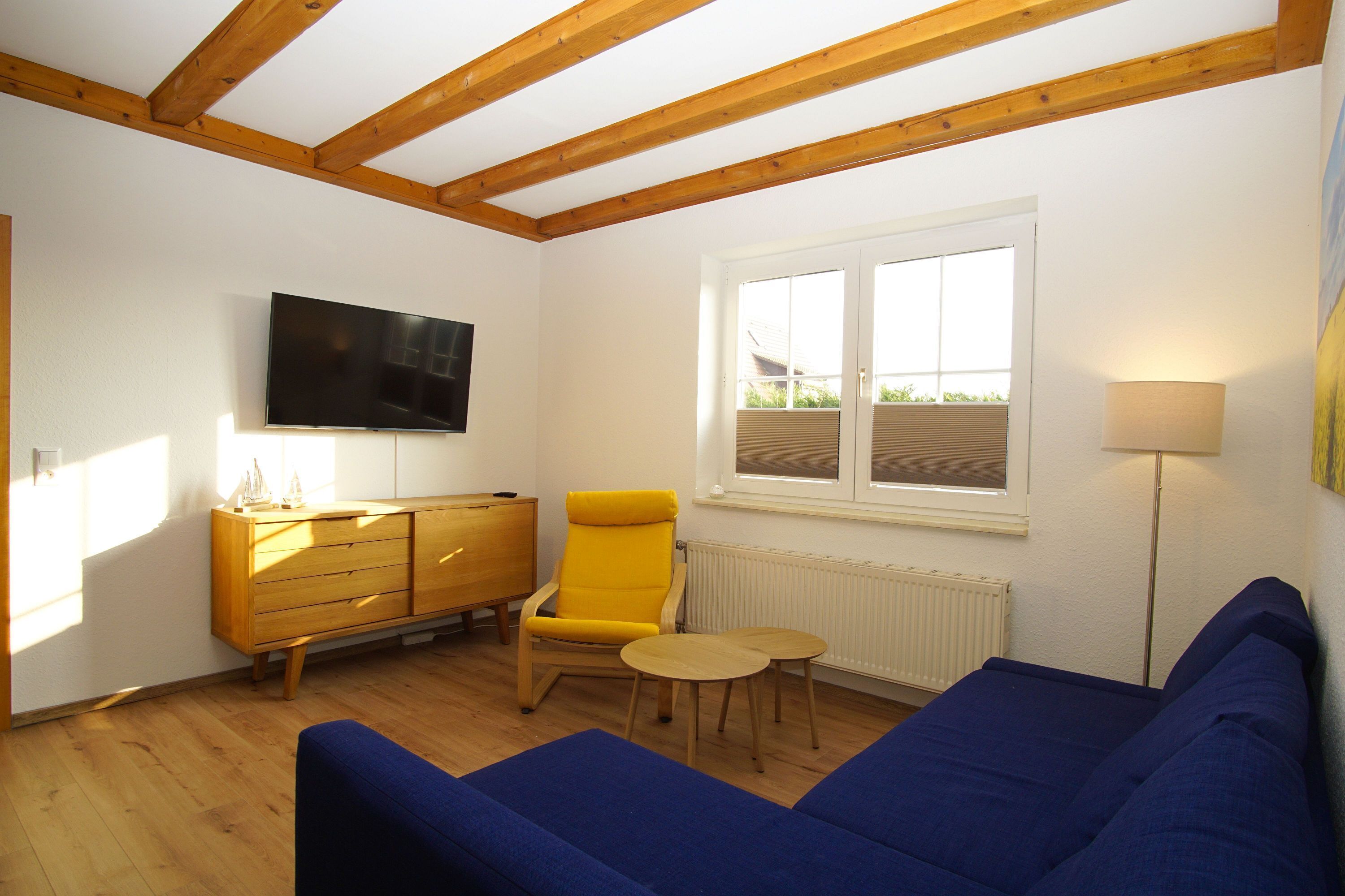 A room with a wooden ceiling, a blue sofa, a yellow chair, a small wooden table, a TV mounted on the wall, and a window with blinds.