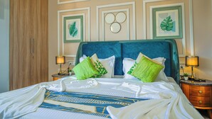 A neatly made bed with a blue headboard, green pillows, and a patterned bedspread. There are two bedside tables with lamps, a wooden wardrobe, and framed artwork on the wall. - Alaya Stays Veera Marina 2BHK (Candolim)