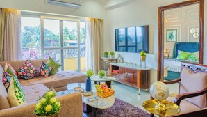 A well-lit living room with a sofa, armchairs, a coffee table, and a TV. There is a balcony with a view of palm trees and a building. - Alaya Stays Veera Marina 2BHK (Candolim)