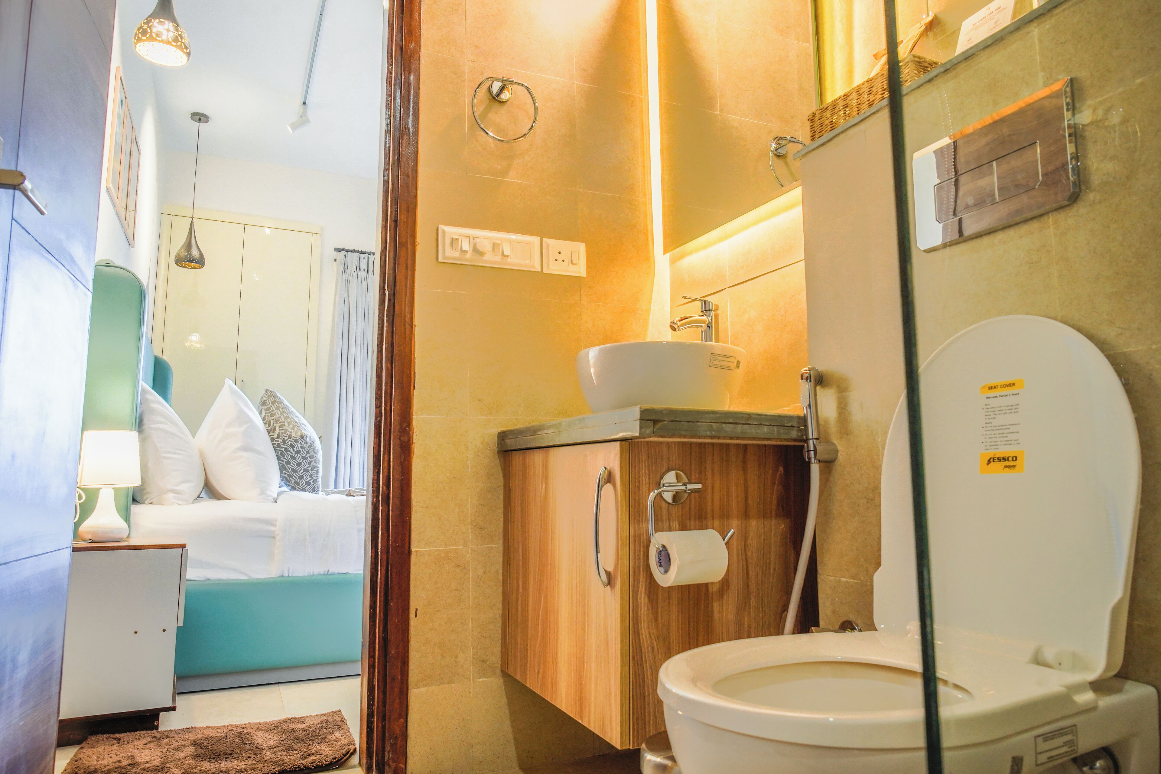 A modern bathroom with a wooden cabinet, a white sink, and a toilet. There is a bed with white bedding and a blue headboard in the adjacent room.