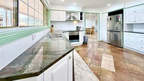 A kitchen with a granite countertop, white cabinets, a stainless steel refrigerator, and a window with blinds.