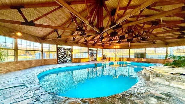 An indoor swimming pool with stone flooring, wooden beams, and large windows.