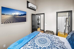 A bedroom with a large bed, a wall-mounted air conditioner, a door leading to a bathroom, and a framed picture on the wall.