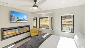 Un dormitorio moderno con una cama grande, chimenea, un sillón amarillo y tres ventanas con vistas panorámicas.