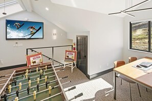 A room with a foosball table, a television displaying a sports event, and a large window with a view.