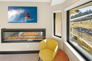 A room with a fireplace, a yellow armchair, a large window with a view of a snowy landscape, and a painting of a skier on a mountain.