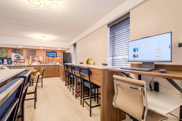 A modern office space with a long counter, multiple chairs, a desktop computer, and a large window with blinds. - Clarion Pointe Winston – Salem Downtown (Winston-Salem)