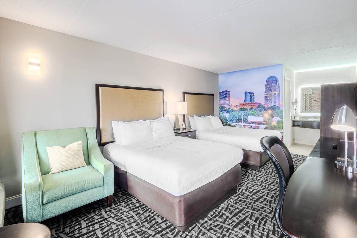 A hotel room with two beds, a green armchair, a dining table, and a cityscape mural on the wall.
