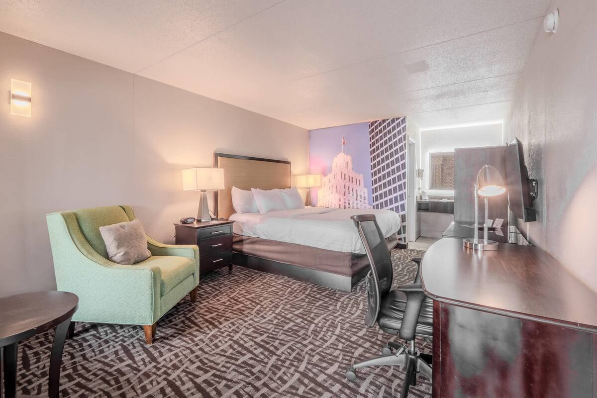 A hotel room with a bed, a desk, a chair, a lamp, and a wall with a cityscape painting.