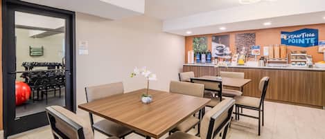 A hotel dining area with a wooden table and chairs, a counter with coffee and snacks, and a fitness center visible through a glass door.
