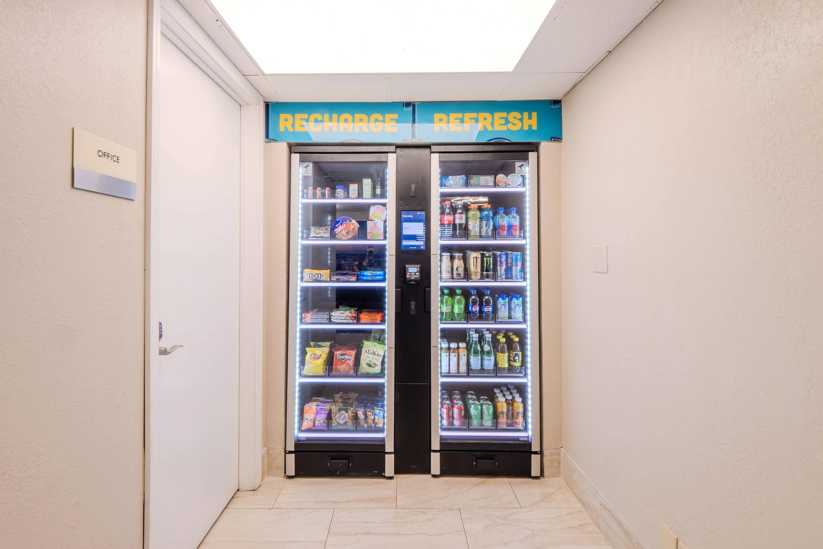 Een gang met twee gekoelde drankautomaten, gelabeld 'RECHARGE' en 'REFRESH', en een bord met de tekst 'OFFICE'.