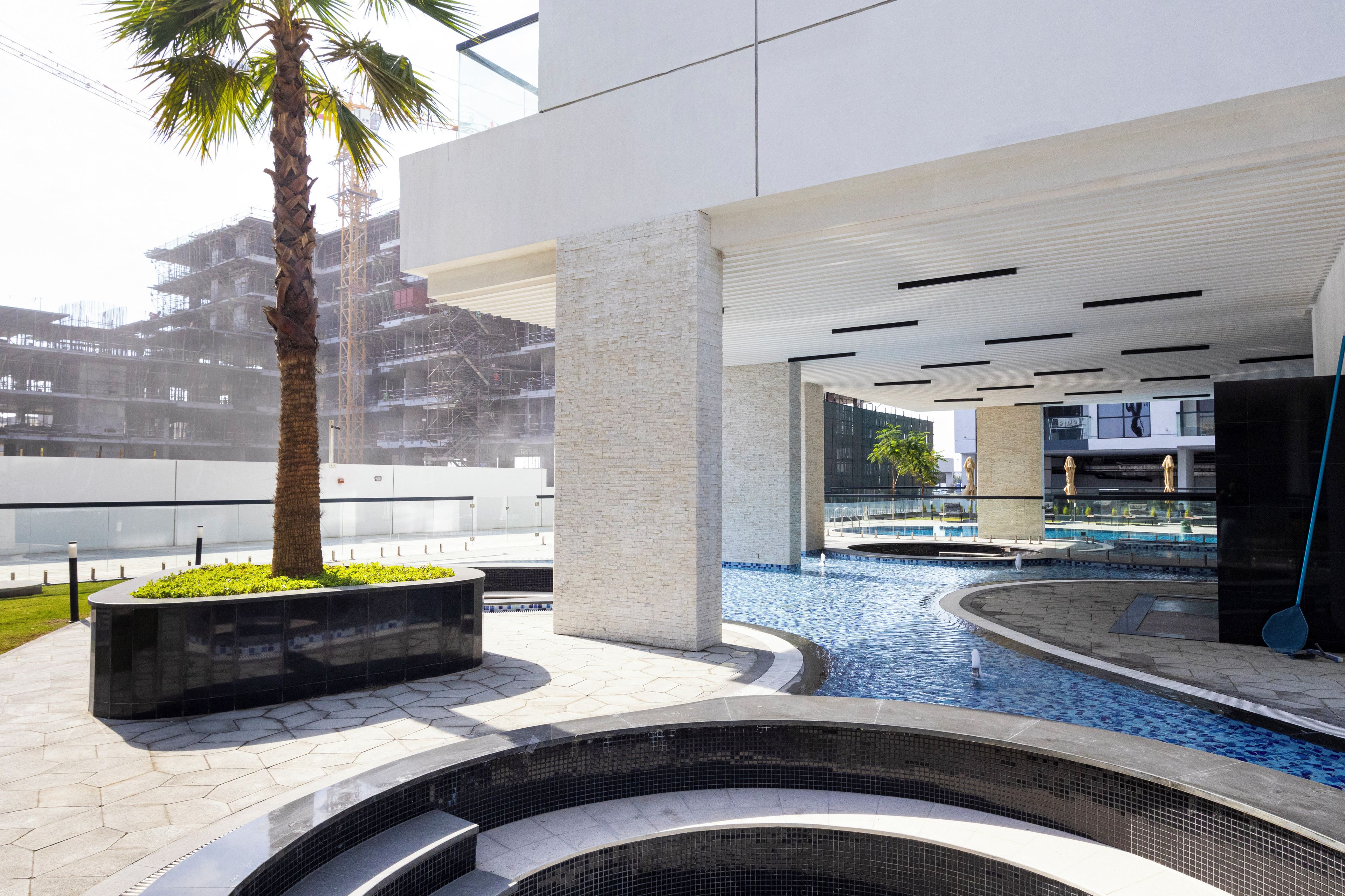A modern building with a swimming pool, palm trees, and a view of a construction site.