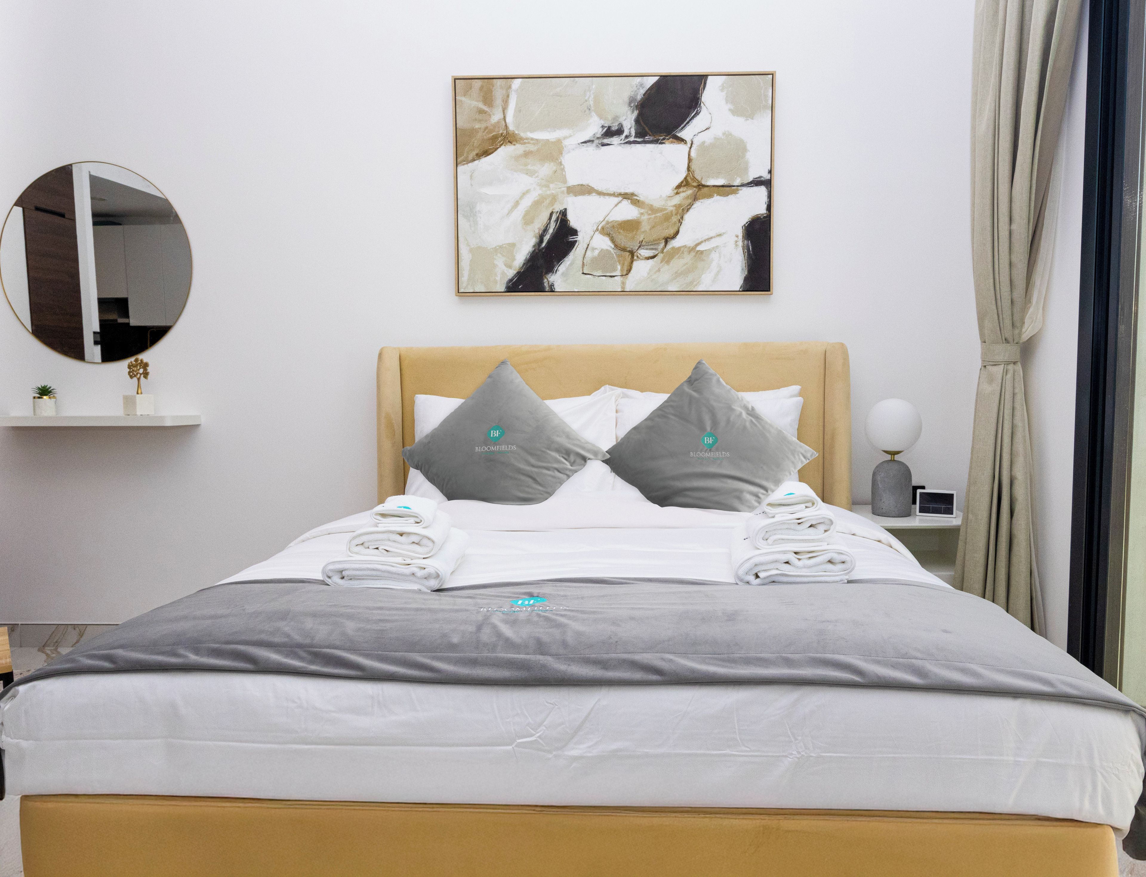 Un lit bien fait, avec une couette grise et blanche, deux oreillers ornés d’un logo, une table de chevet avec une lampe, un tableau abstrait accroché au mur, un miroir rond et un rideau.