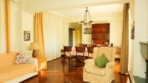 Una sala de estar con sofá, sillón, mesa de comedor y sillas. Se observa una araña de techo y una chimenea.