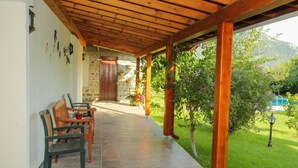 Un patio techado con muro de piedra, sillas de madera, piso de baldosas y vista a un jardín y una piscina.