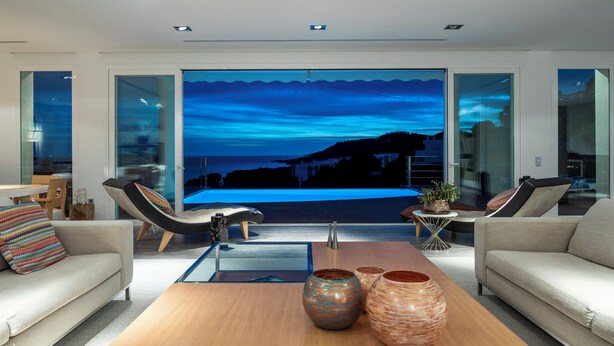 A modern living room with a large glass door leading to a balcony with a view of the sea. There are two sofas, a coffee table with decorative vases, and a small side table with a plant.