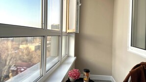 A corner room with a view of the city, a small table with a cup and flowers, and a chair.