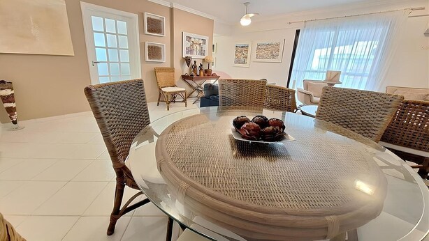 A dining area with a round glass table surrounded by wicker chairs, a bowl of fruit on the table, and a view of the city through large windows.