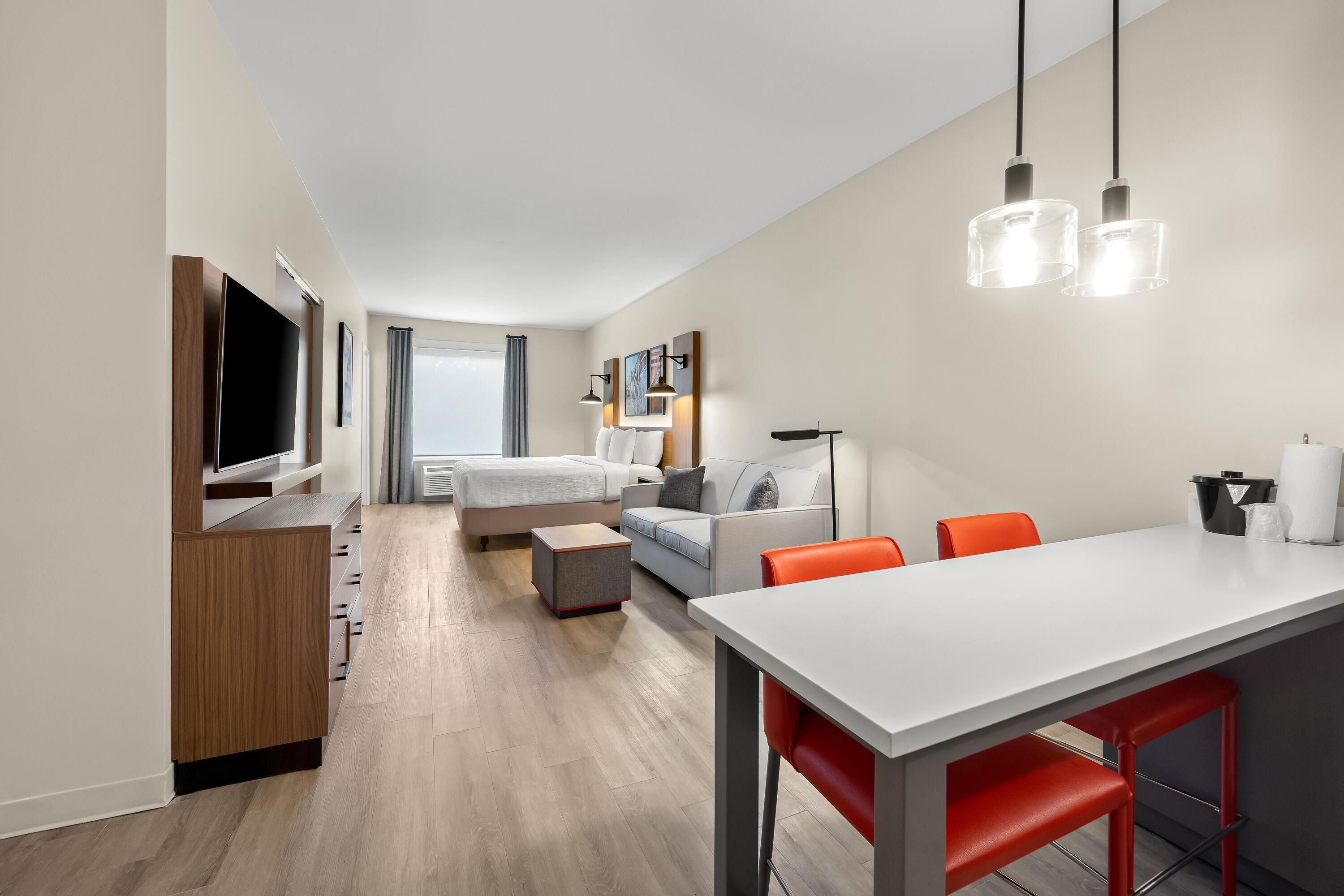 A modern hotel room with a flat-screen TV, a bed, a sofa, a dining table with chairs, and a large window.