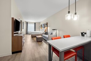 A modern hotel room with a flat-screen TV, a bed, a sofa, a dining table with chairs, and a large window. - Hawthorn Extended Stay by Wyndham Mebane (Mebane)