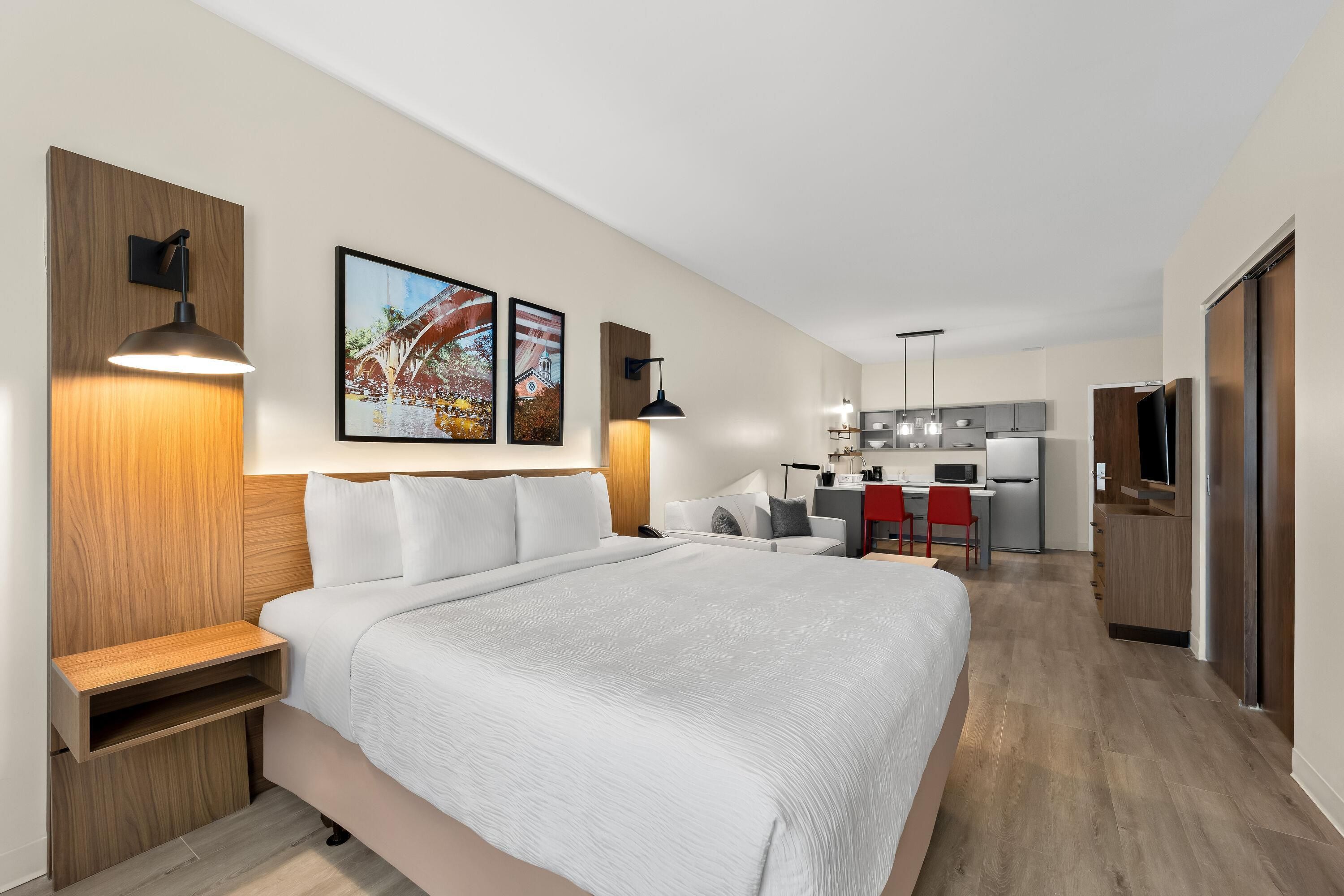 A modern hotel room with a large bed, bedside tables, a desk, and a kitchenette.