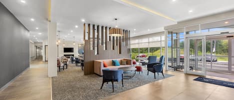 A modern hotel lobby with a seating area, a fireplace, and a large window offering a view of the outdoors.