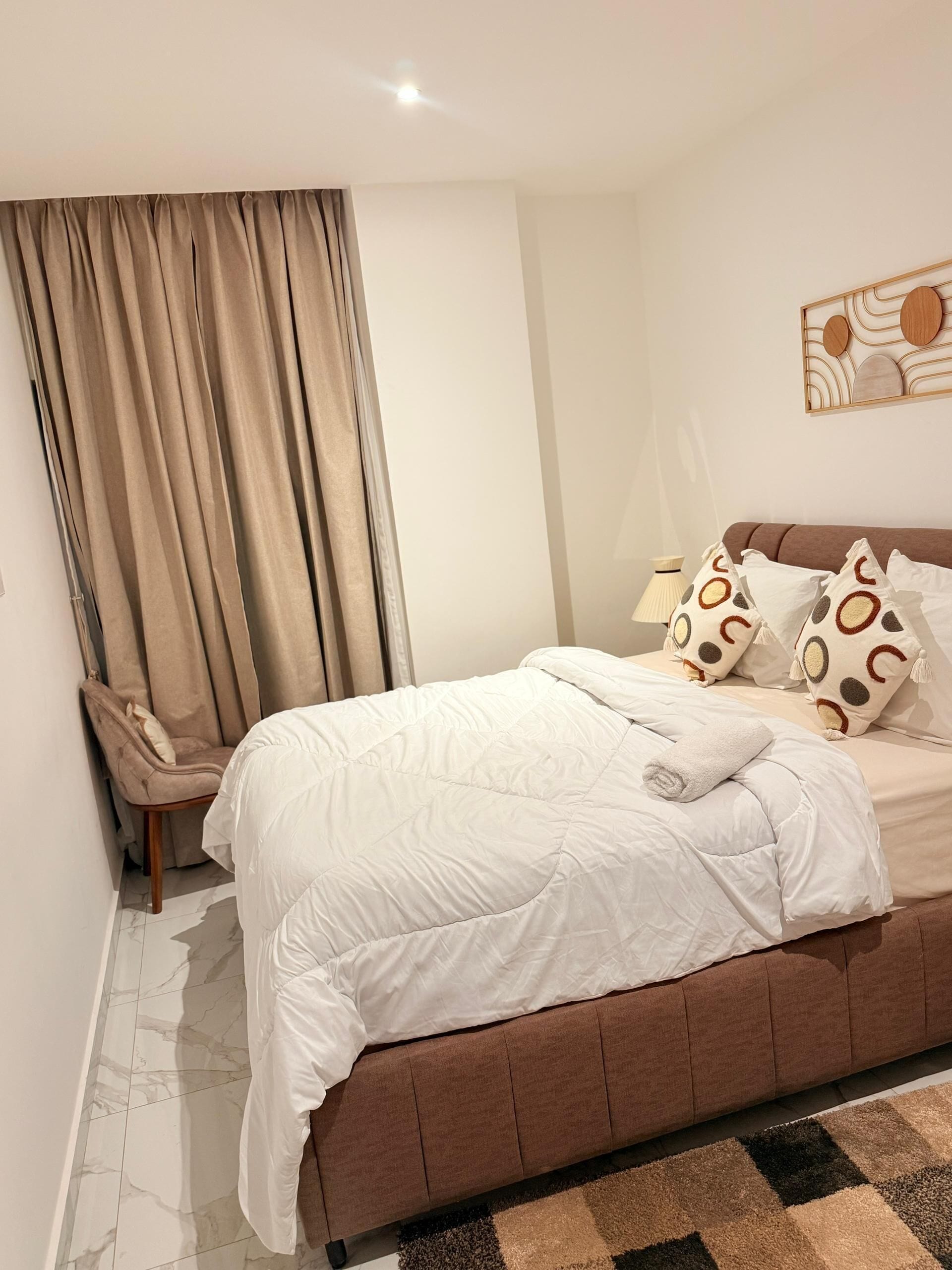 A bedroom with a bed, curtains, a chair, a lamp, and a wall art piece.