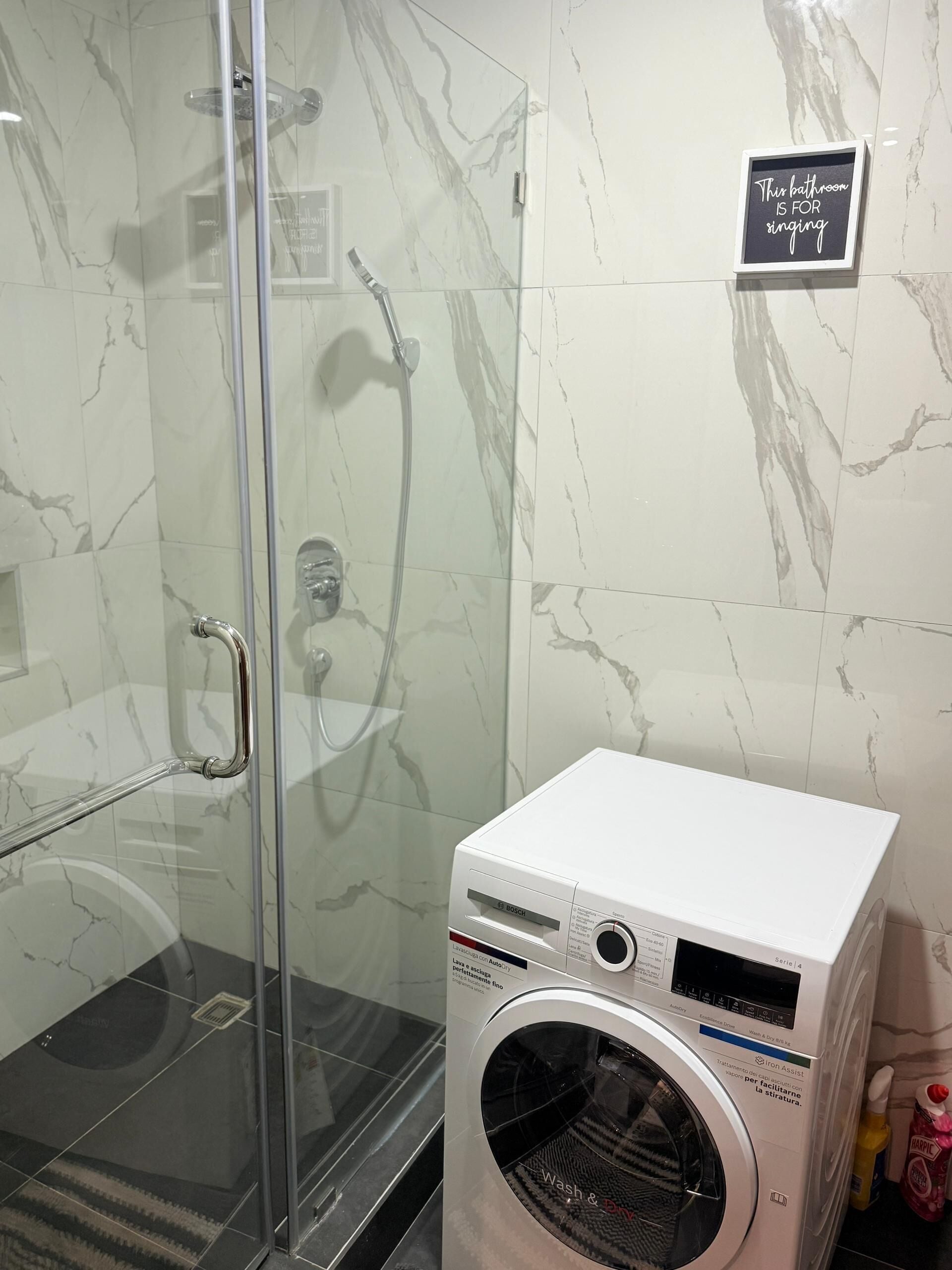 A bathroom with a glass shower enclosure, a Bosch washing machine, and a sign stating 'This bathroom is for singing.'