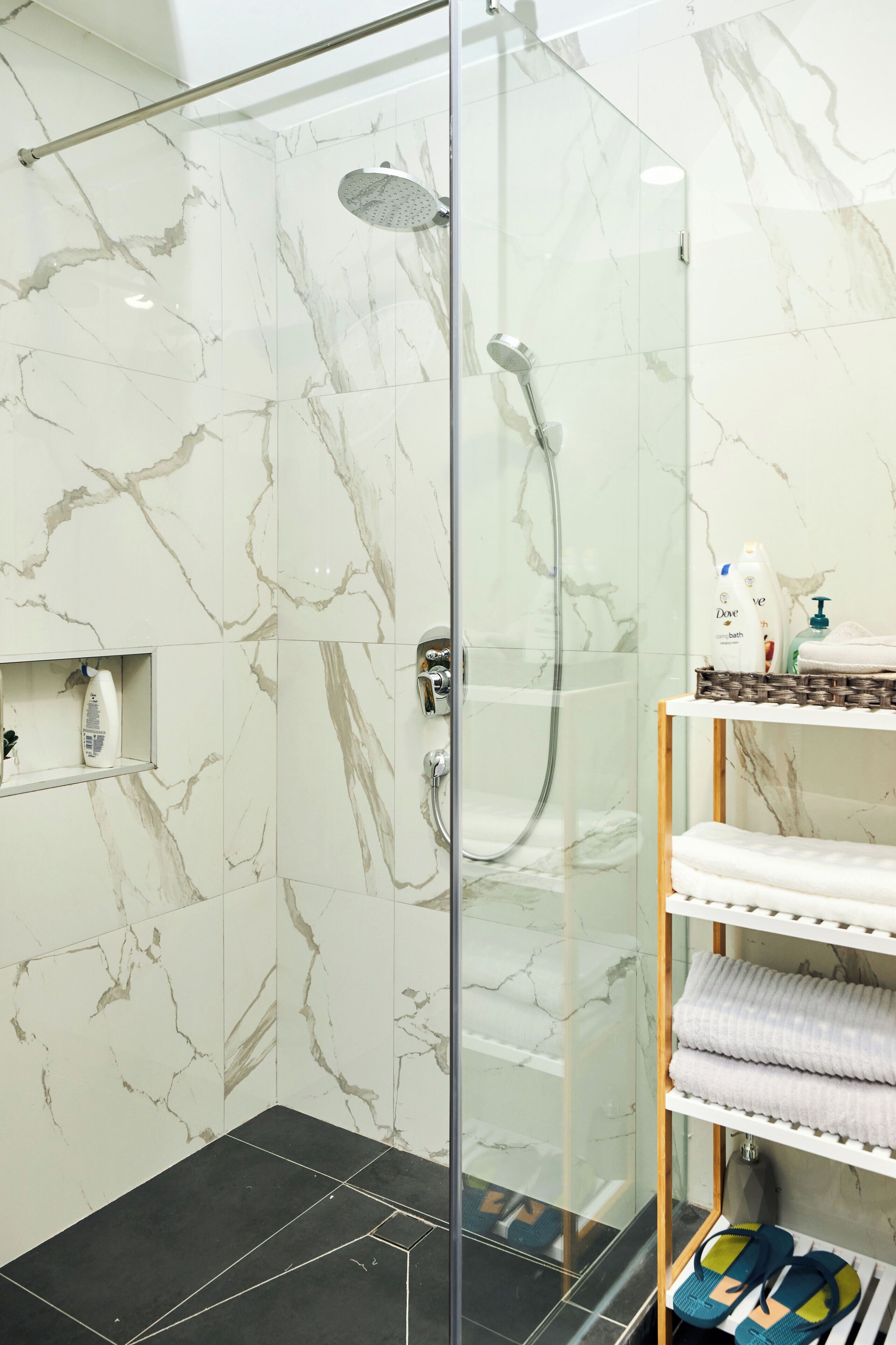 Ducha con mampara de vidrio, cabezal de ducha manual, repisa con artículos de aseo y toallero con toallas dobladas.