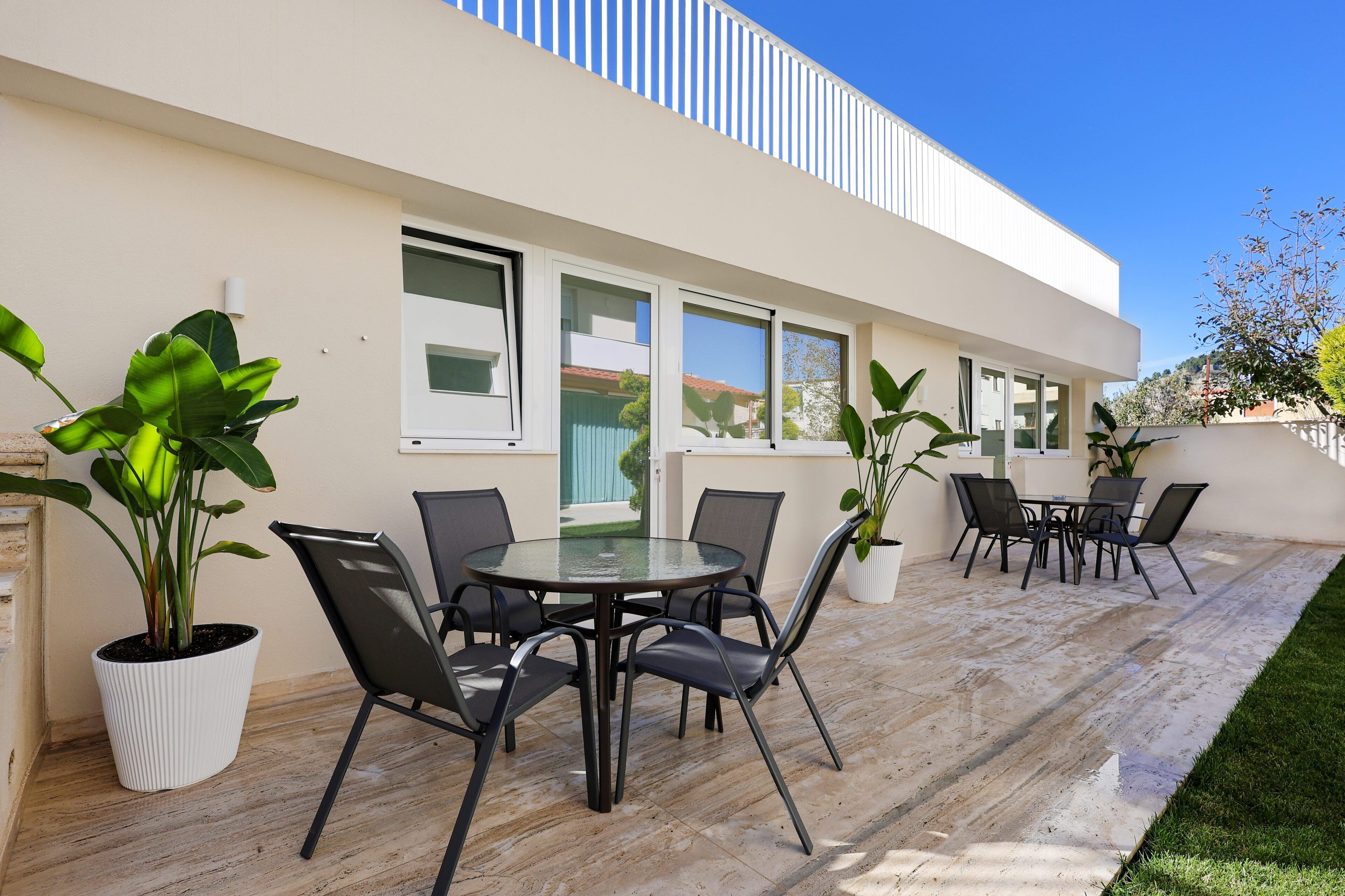 A modern outdoor patio with a round glass table and black chairs, surrounded by potted plants.