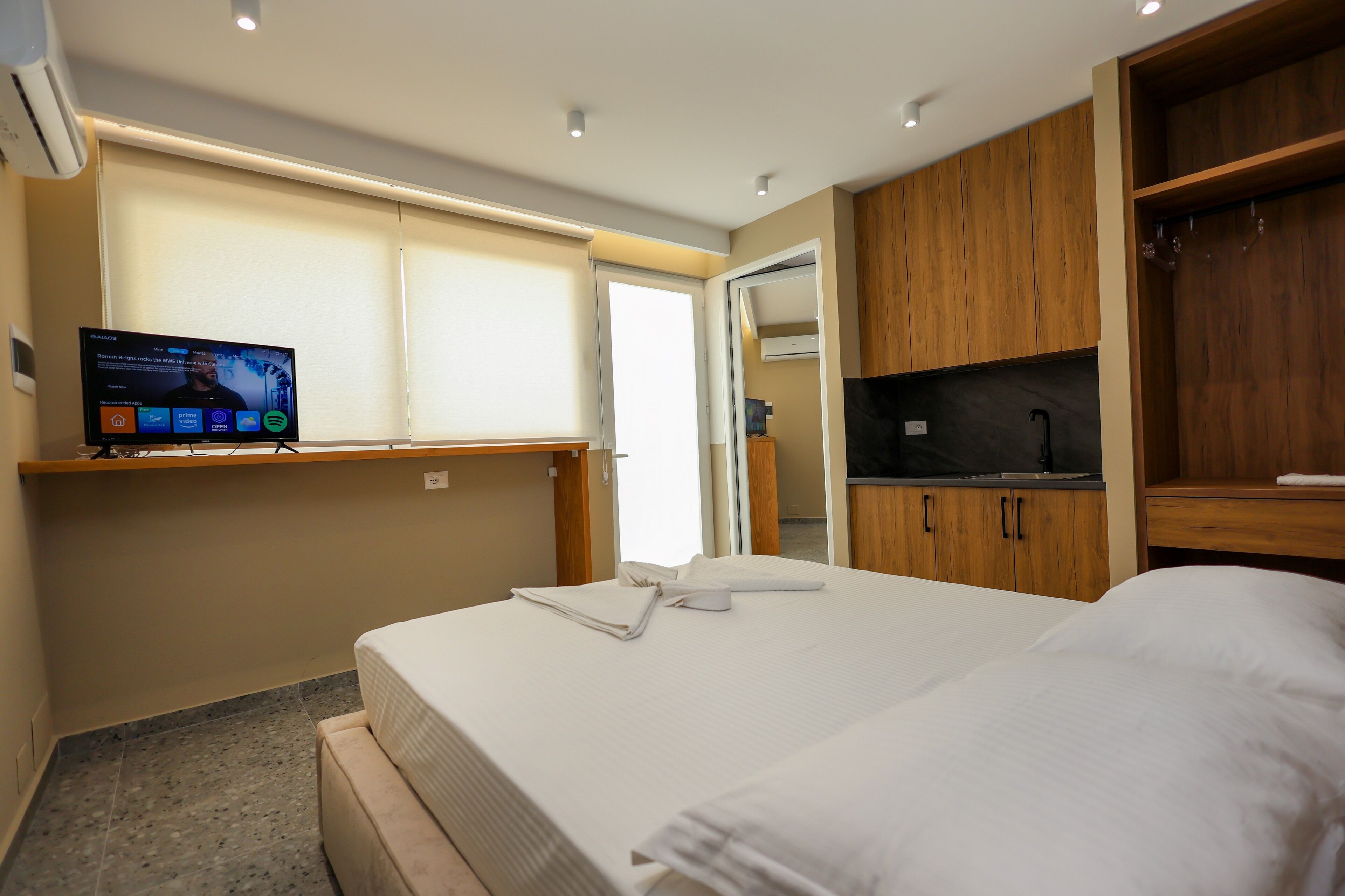 Una habitación de hotel con una cama, un televisor en un soporte, armarios de madera y una ventana con persianas.