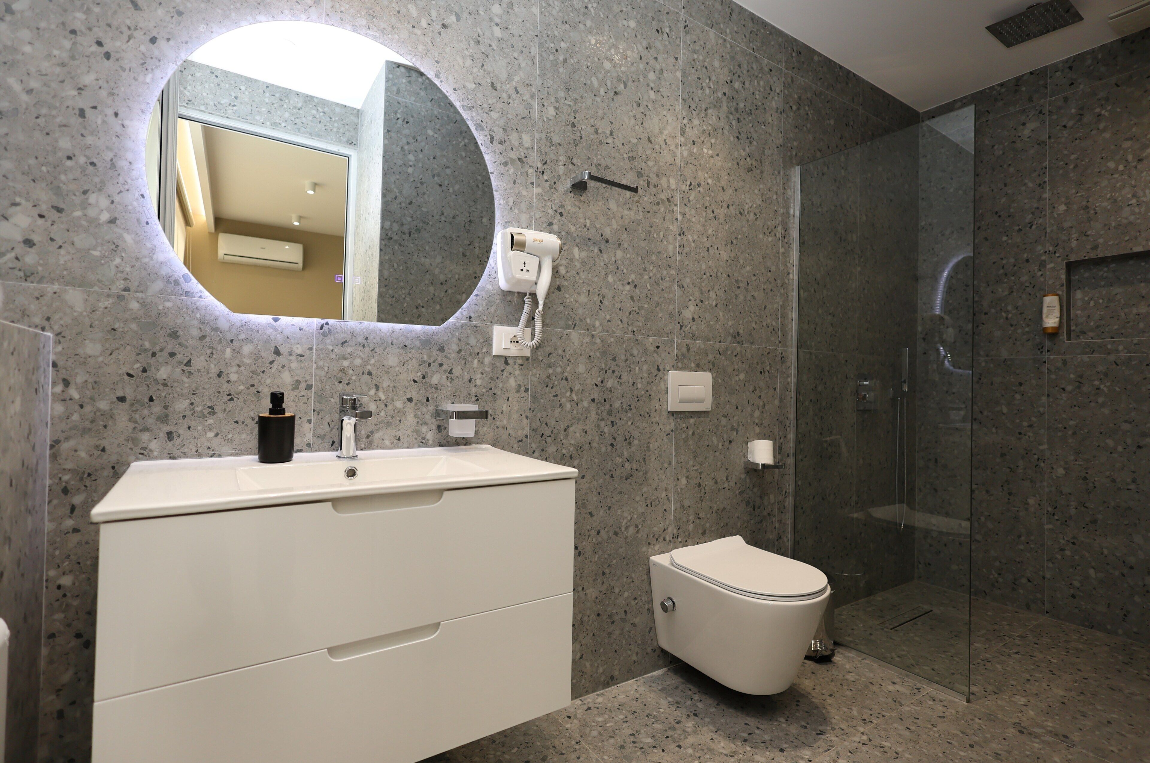 Un baño moderno con encimera de granito, un lavabo blanco, un espejo redondo, inodoro y ducha con mampara de vidrio.
